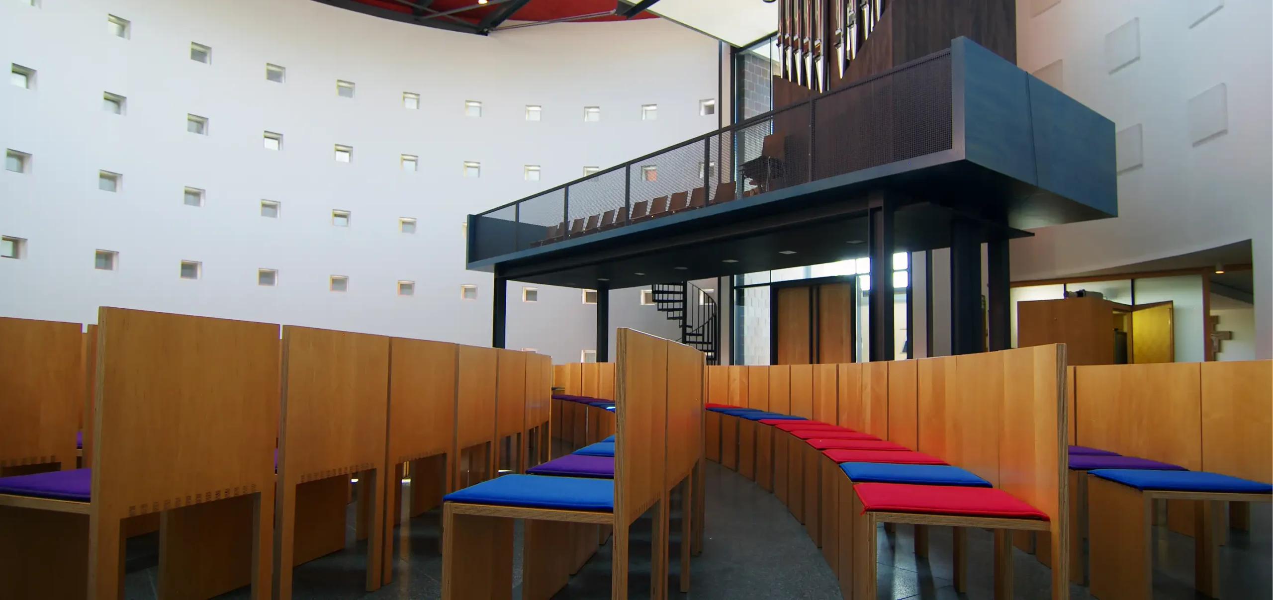 Kirche St. Klara in Ulm, Innenraum, Kirchenschiff, Gebetsraum, Orgelamphore, kreisförmige Anordnung der Bestuhlung, Holzstühle mit Fingerzinken, blaue, rote und lila farbige Sitzkissen