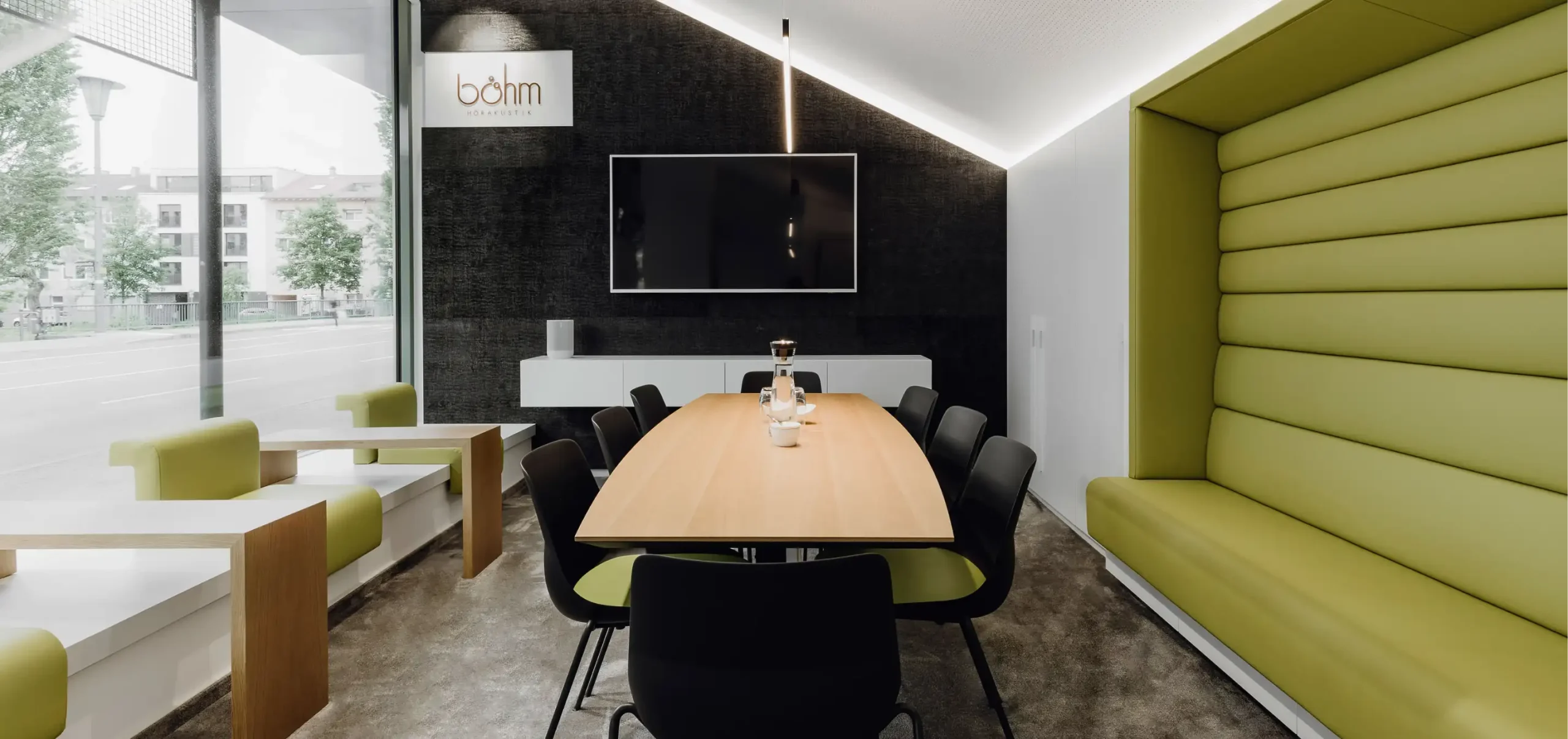 Büroflächen Böhm, andere Perspektive mit Fenster zur Straße, zur linken Sitze mit Beistelltisch, Abgeflammte, gerissene Holzverkleidung in Schwarz mit indirekter Beleuchtung und Monitor, Konferenztisch mit Stühlen, Sitznische