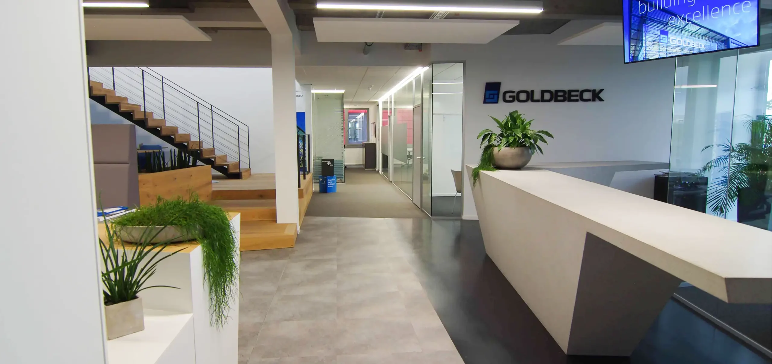 Innenausbau der Büroräume Goldbeck Eingangsbereich, gespachtelter Betonkubus links als Empfangstheke, Wand mit Goldbeck Logo, hinten Büros durch Glaswände getrennt, links Holz- Stahltreppe