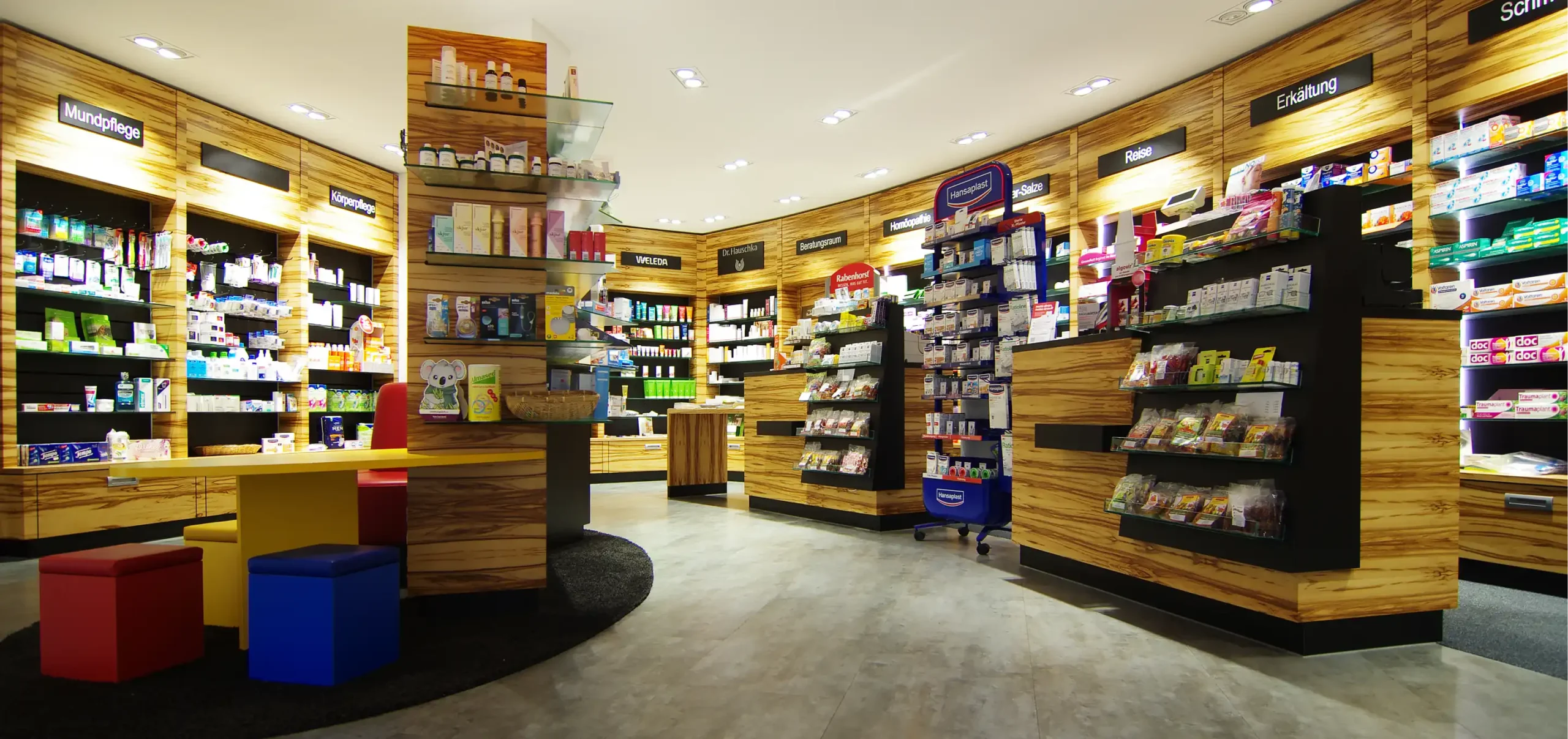 Andere Perspektive des Verkaufsraum der Tal Apotheke, Warte und Sitzgelegenheiten, Verkaufstresen, mit Wandschränke aus Holz und individuellen Regalsystemen für die Verkaufsfläche