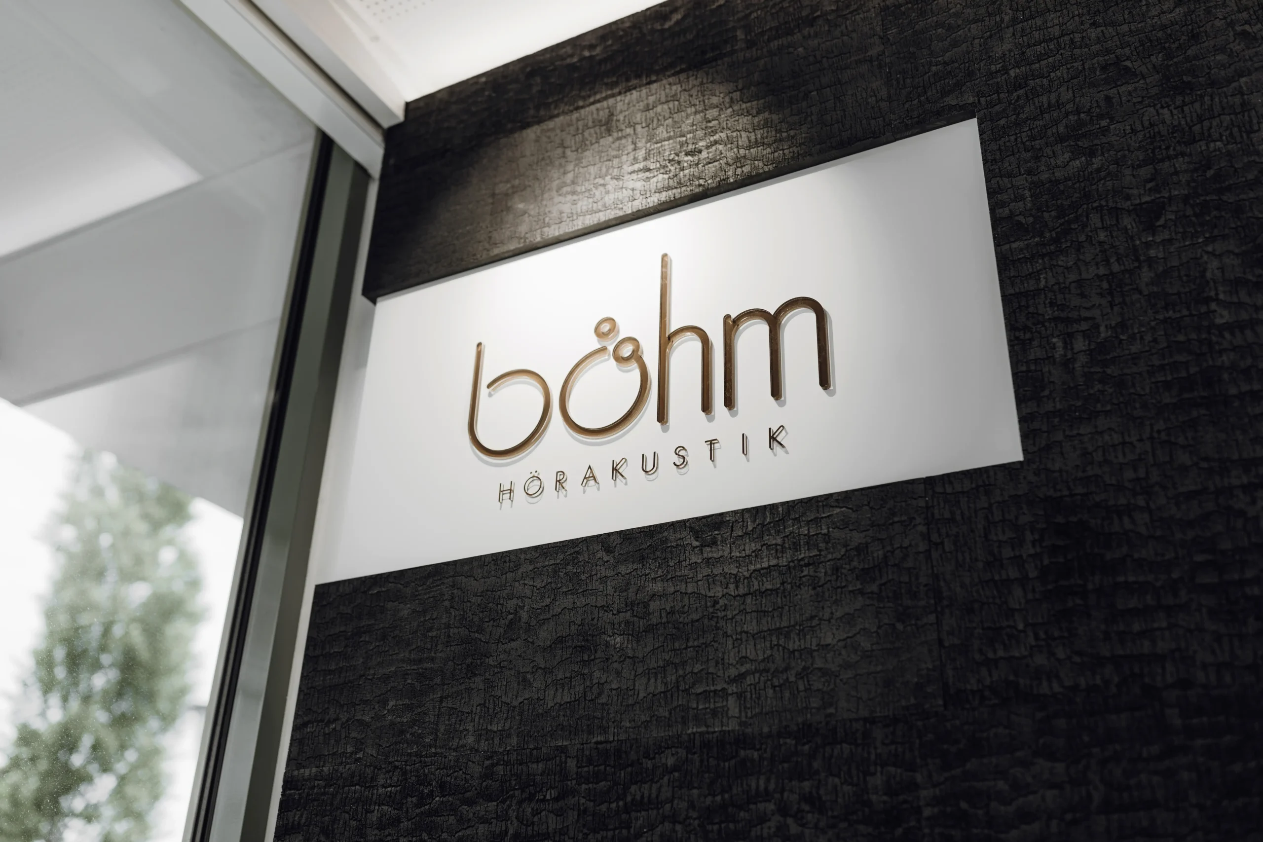 Büroflächen Böhm, Detailaufnahme von abgeflammten und gerissene Holzverkleidung in Schwarz mit indirekter Beleuchtung und integrierten Firmen Logo von Böhm Hörakustik 