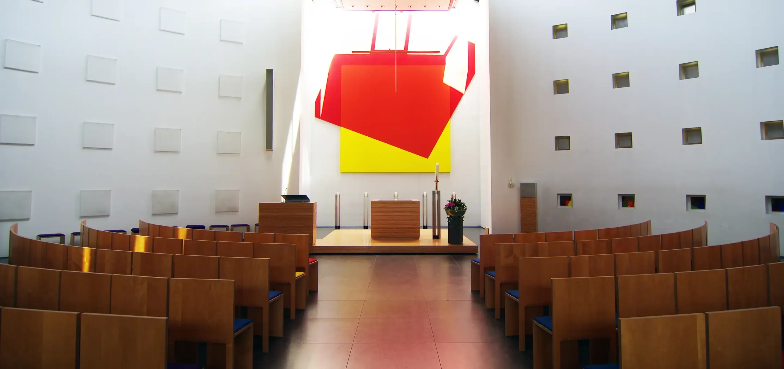 Kirche St. Klara in Ulm, Kirchenschiff in zentral Perspektive, Weise Wände, schlichter Holzaltar leicht erhöht auf einen Holzpodest, indirekte Tageslicht Beleuchtung der Altar Nische, abstrakte Kunstdekoration in der Altarnische