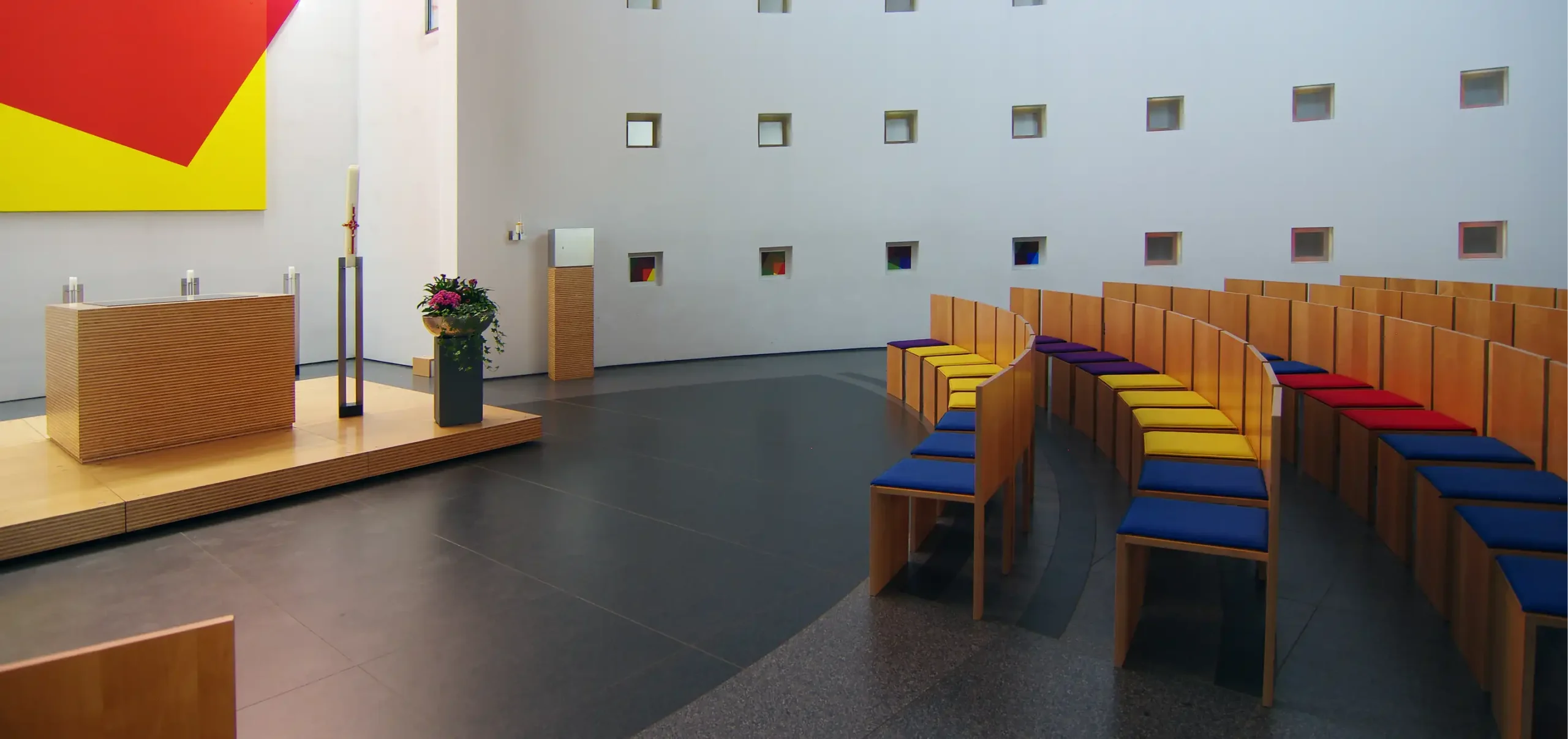 Kirche St. Klara in Ulm, Seitenansicht, Kirchenschiff, ovale Anordnung der Bestuhlung, Holzstühle mit Fingerzinken, blaue, rote, gelbe und lila farbige Sitzkissen, Holzaltar auf niedriger Amphore aus Holz, schlichte Kerzenständer mit Kerzen