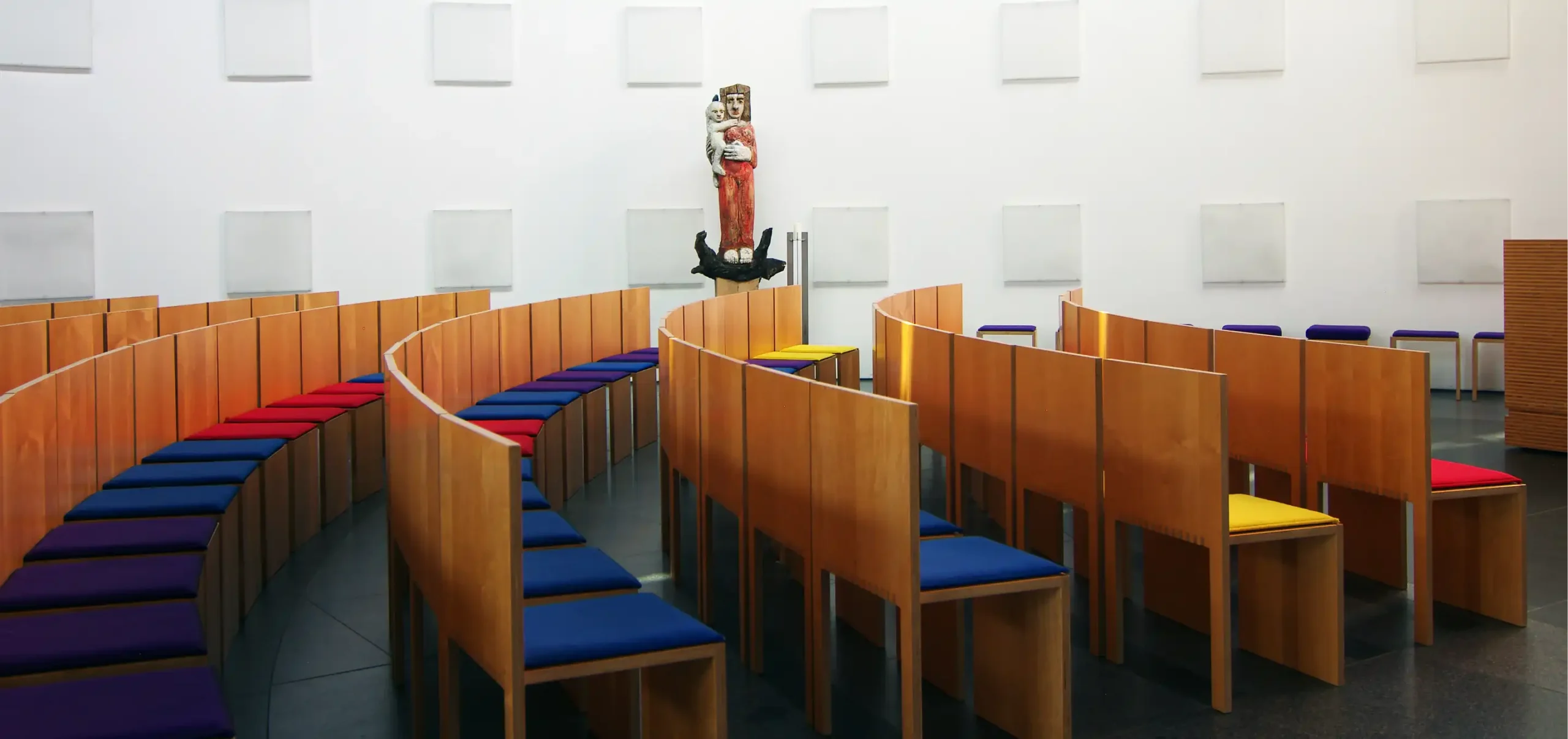 Kirche St. Klara in Ulm, Kirchenschiff, abstrakte Marienstatue im Hintergrund, kreisförmige Anordnung der Bestuhlung, Holzstühle mit Fingerzinken, blaue, rote und lila Sitzkissen