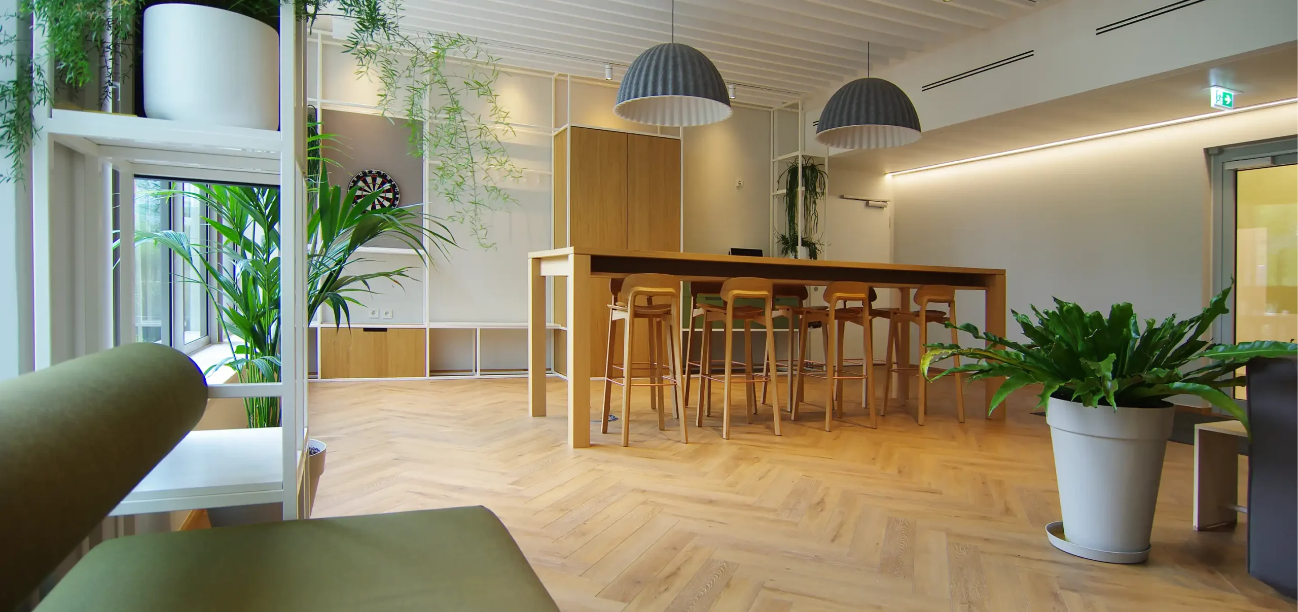 Büroräume von Bloom Offices, Fenster, grünes Polstermöbelstück, Hochtisch aus Holz mit Bestuhlung, Hängelampen grau weiß, weiße Stahlregale und frische Grüntöne, Pflanzen