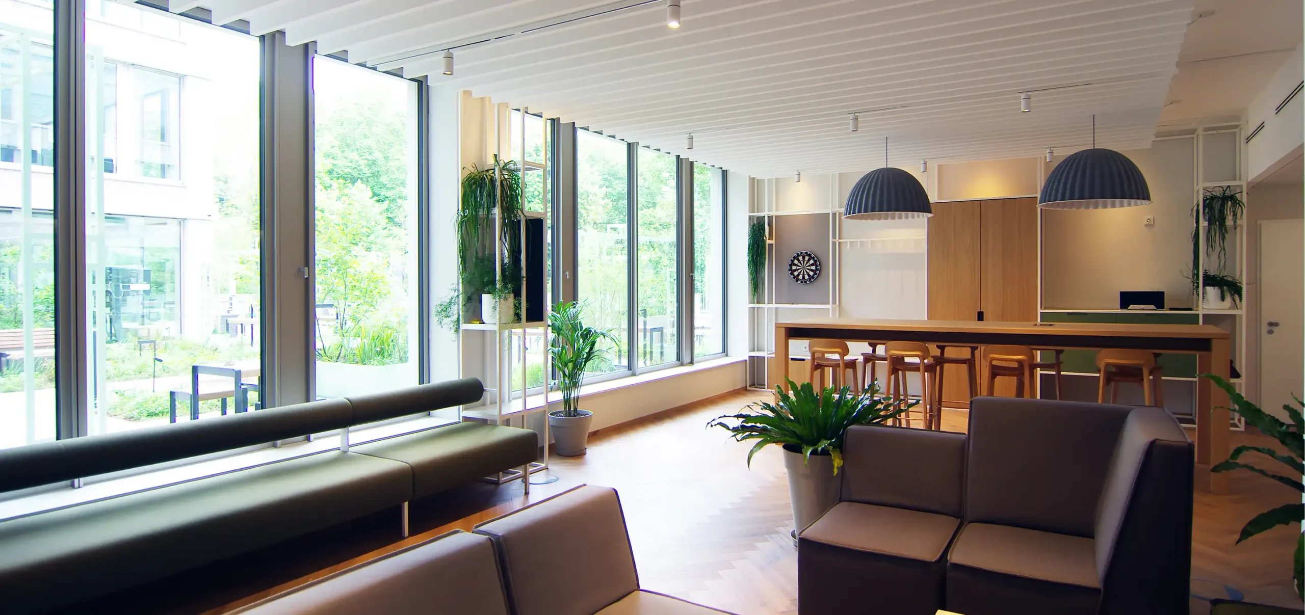 Büroräume von Bloom Offices, Fenster, Sitzbereich mit Polstermöbel, Hochtisch aus Holz mit Bestuhlung, Hängelampen grau weiß, weiße Stahlregale und frische Grüntöne