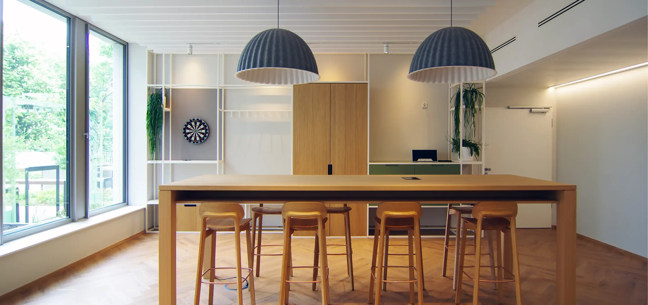 Büroräume von Bloom Offices, Fenster, Hochtisch aus Holz mit Bestuhlung, Hängelampen grau weiß, weiße Stahlregale und frische Grüntöne
