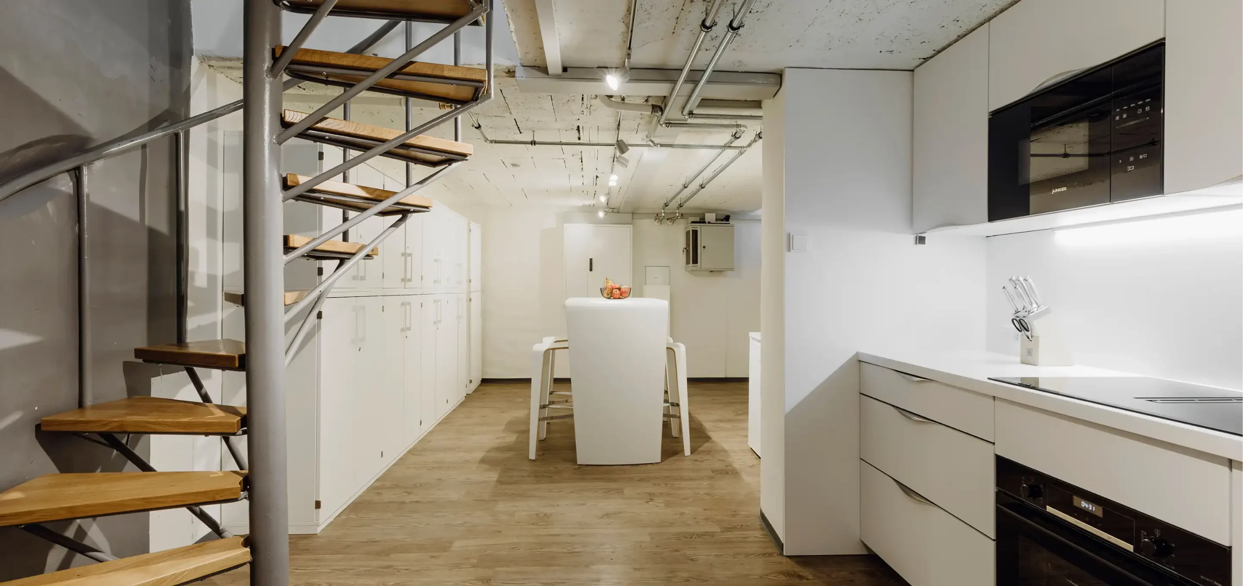 Büroflächen Böhm, Raumkonzept vom Kellergeschoss, Küche und Küchenmöbel in weiß, Wendeltreppe aus Holz und Stahl, Tisch mit Hochstühlen, Boden mit Holzdielen