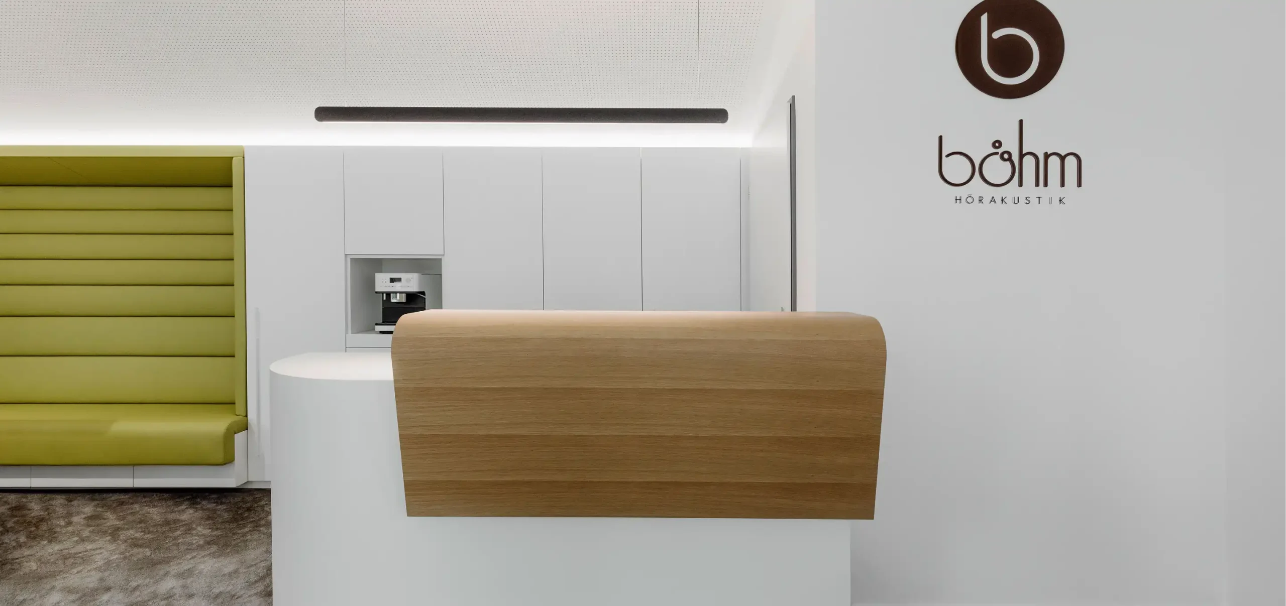 Büroflächen Böhm, weiße Theke lackiert mit Holzbestandteilen, rechts Wand mit Böhm Hörakustik Logo, weißer Wandschrank mit Nische für Barista-Espressomaschine, grüne gepolsterte Sitznische, Hängelampe