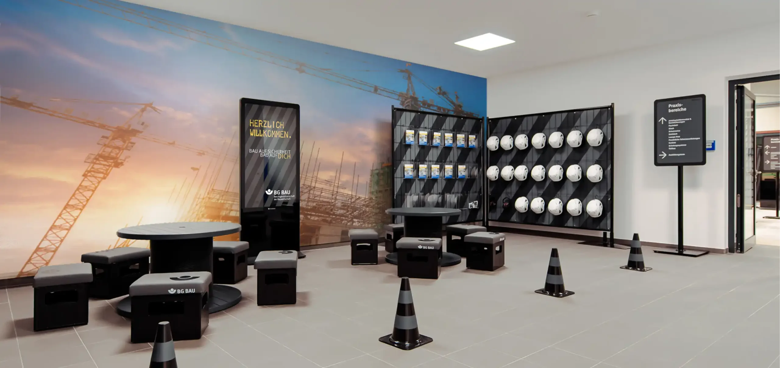 Ausstellungsraum BG-Bau, Sitzgelegenheit, Warnhütchen, Kabelrollentisch, Werbetafel, Text: Herzlich Willkommen, Bau auf Sicherheit Bau auf Dich, Broschürenstellwand, Hinweisschild