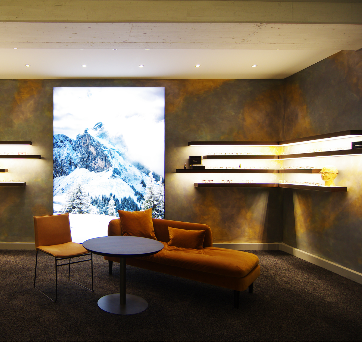 Verkaufsräumlichkeiten, an der Wand ein Lichtpaneel, Regale mit indirekter Beleuchtung für die Produkte, Produkte sind Brillen, Sofa, Tisch, und Stuhl, braun und orange Farbtöne