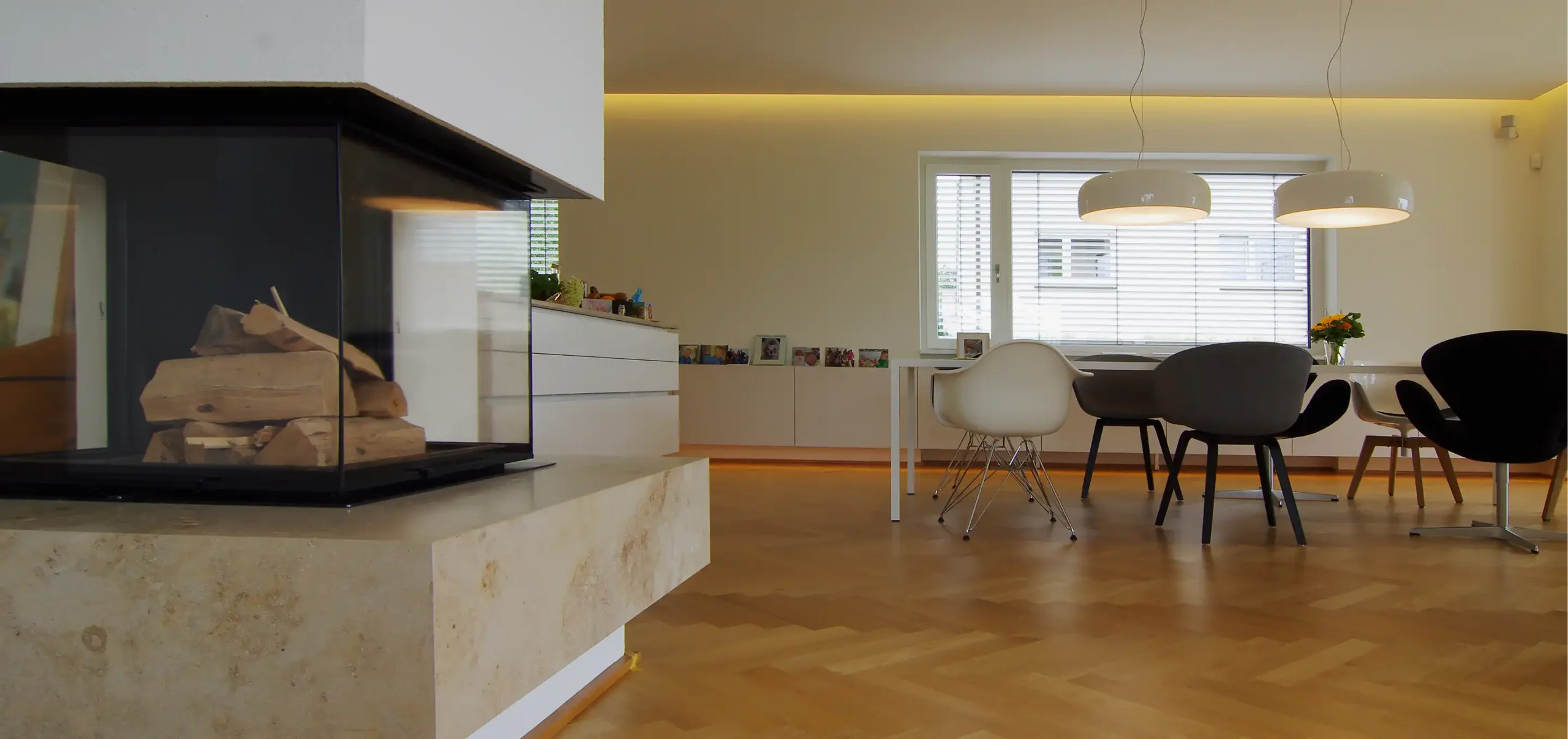 Wohnzimmerbereich mit Kamin, Tisch und unterschiedliche Designer - Stühle, Hängelampen über den Tisch, weiße Sideboards, indirekte Wandbeleuchtung, Ausführung des Gewerks Miller Ladenbau