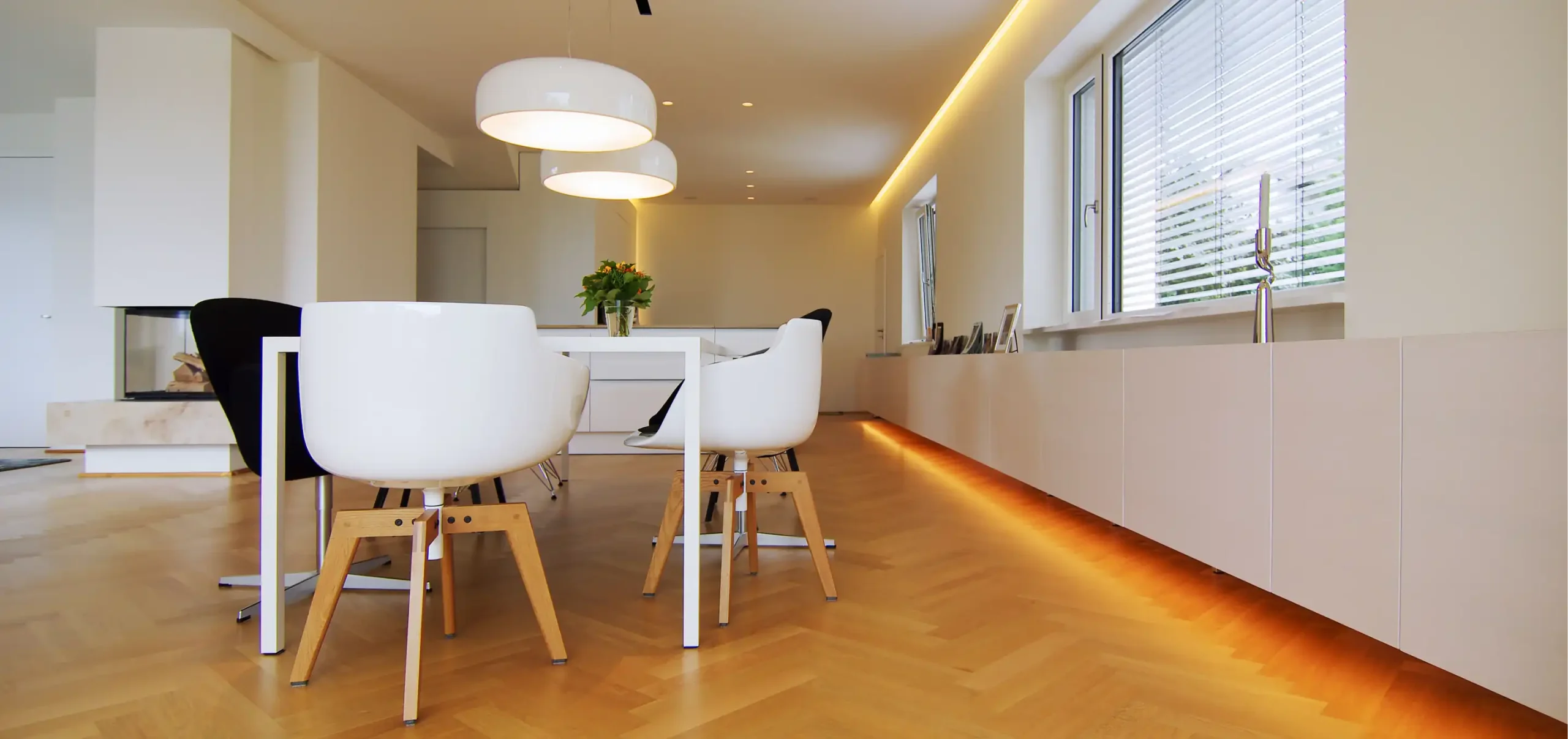 Wohnzimmer, mit Tisch und Designer Stühlen, zwei Designer Lampen an der Decke, durchgängige Wandverkleidung unter dem Fensterbereich, mit indirekter Bodenbeleuchtung