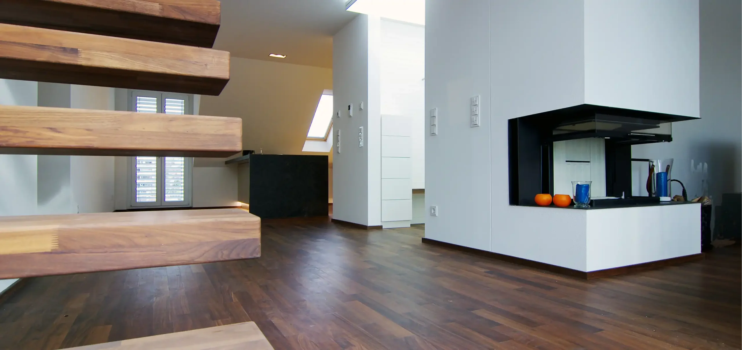 Maisonette-Wohnung mit Dachschräge, Wohnzimmerbereich mit Kamin, Treppe aus Holz ohne Geländer, handgefertigte Möbel, dunkle Holz-Dielen, Hintergrund Küchenzeile